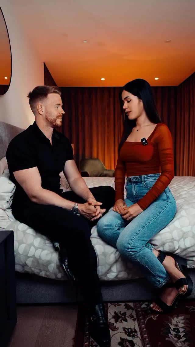 A seated man and woman face each other on a bed, both looking towards each other, suggesting a close relationship. The man has ginger hair and is wearing a black top, while the woman has dark hair and wears a red top and jeans. The background hints at a neutral setting. Warm lighting adds to the intimate feel of the scene.