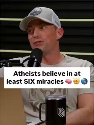 A man in a grey cap and shirt speaks into a microphone. Above him, a text box reads, 'Atheists believe in at least SIX miracles'. A black mug with a logo sits on a table in front.