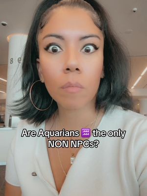 A selfie-style shot of a woman with dark hair and prominent dark eyes in front of a blurred background. She wears a white top and hoop earrings. Text overlay at the bottom asks "Are Aquarians the only NON NPCs?" The woman appears surprised, with wide eyes and a slightly pursed mouth.