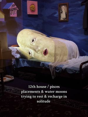 An image depicting a surreal bedroom scene. In the foreground, a large, illuminated, human-like face rests on a bed, its eyes closed. The face appears to be part of a giant head, positioned as if it's resting. In the background, there are elements of a bedroom, like wall art, a bed frame, and a nightstand. The color palette is predominantly muted blues and yellows, creating a dreamlike atmosphere. Text overlaid on the image describes the scene.