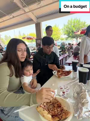 A family is seated at an outdoor table, eating pizza. A woman holds a baby in her lap. There are two other people at the table eating pizza. Text overlayed at the top.