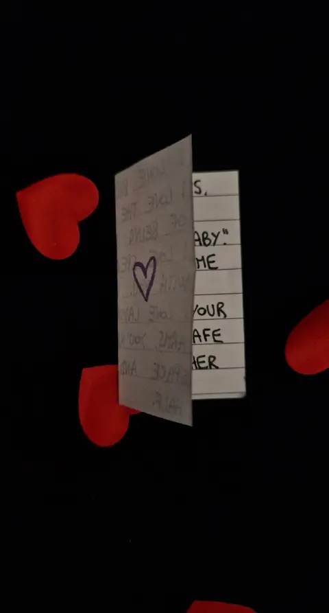 A photograph shows a partially open, folded card against a black background with scattered red heart-shaped confetti. The card is turned to the side, revealing handwritten text and a drawn purple heart.
