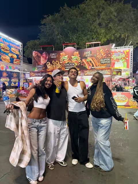 Image captures a group of four young adults posing together at an outdoor food festival at night. A variety of food vendors are visible in the background with bright lights and signs. The group is diverse, dressed in casual, late 90s fashion, and smiling. They are positioned in front of the food stalls. The composition is well-lit with strong colors. The overhead lighting casts some shadows, giving depth.