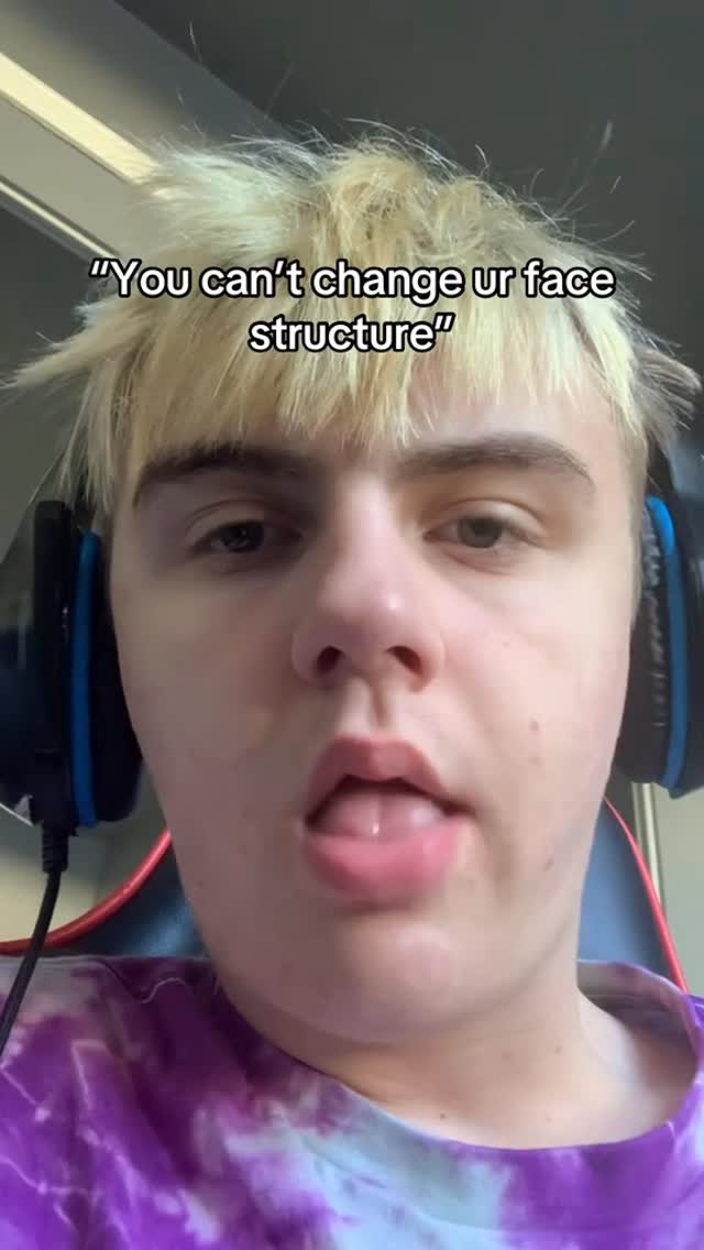 A selfie-style shot featuring a young man with styled, light-colored hair and a purple-and-white tie-dye shirt. He's wearing headphones and has a slightly open mouth, with his tongue visible. The background is a dark, blurred interior.