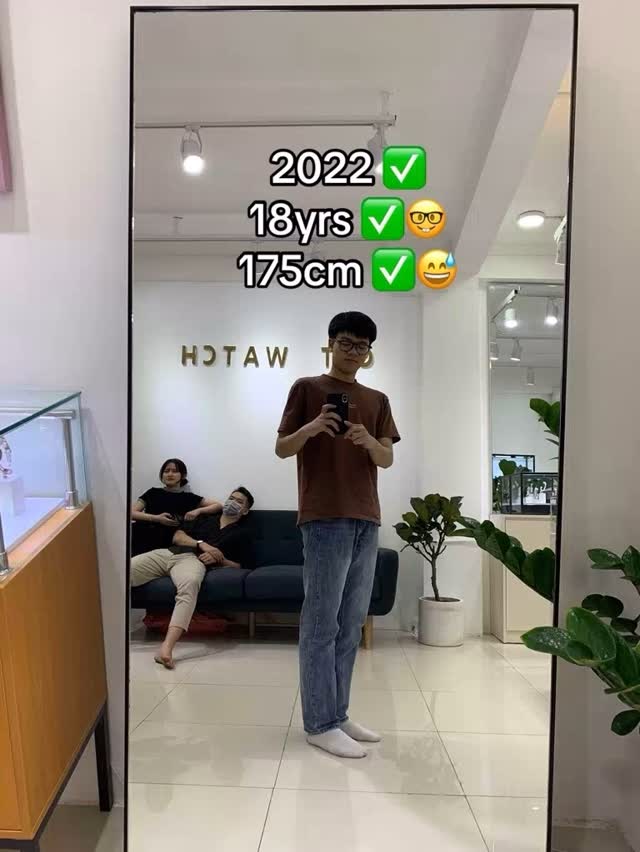 A mirror reflects a young man standing, holding a phone, in front of a retail shop setup with two seated people on the background. Text overlays highlight the year, his age, and reported height.