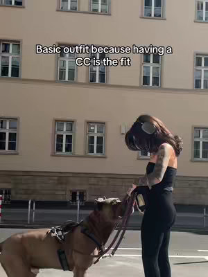 A woman in all-black athletic wear stands in front of a building, holding a leash attached to a large dog. The dog is wearing a harness. Text overlay reads: 'Basic outfit because having a CC is the fit.' The background includes a building with windows and a clear sky.