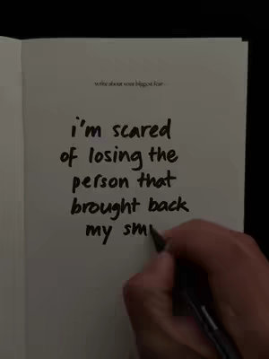 A hand holding a black pen is writing on a page open in a book. The page contains text: 'i'm scared of losing the person that brought back my smi'. The edge of the book is visible on the left and black background color is behind the book pages.