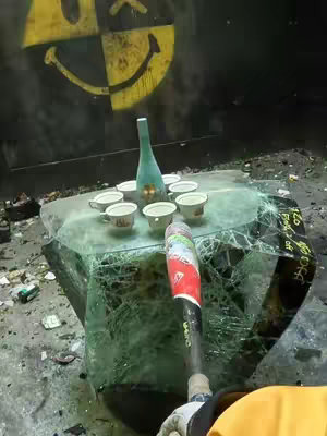 The image shows a point-of-view shot with a baseball bat in the foreground and a shattered glass table in the center holding a turquoise bottle and several white cups, possibly liquor. The background features a black wall with a half-yellow smiley face logo. The overall scene suggests destruction or a release of frustration.