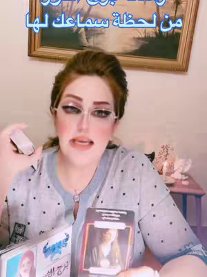 A woman with makeup and eyeglasses is holding tarot cards. The background features a framed picture. The image is a medium shot with a soft, warm lighting.