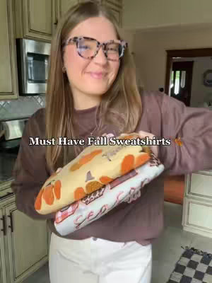 A woman with long, blonde hair and glasses smiles warmly while holding two folded sweatshirts. The image is taken in what appears to be a kitchen, with cabinets and a microwave visible in the background. The sweatshirts have fall-themed prints like pumpkins.  Text overlayed on the image says 'Must Have Fall Sweatshirts'.