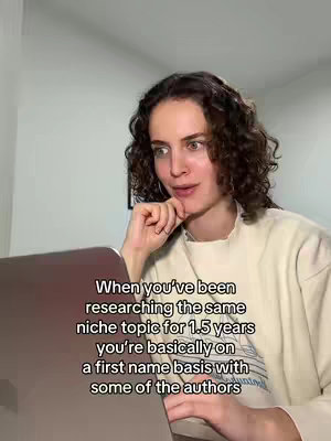 A woman with curly brown hair is seated, looking at a laptop. Text overlayed on the image reads, 'When you've been researching the same niche topic for 1.5 years you're basically on a first name basis with some of the authors'. The background is a plain interior.