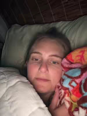 A close-up shot of a woman lying in bed, wrapped in a blanket. The woman has light skin and fair hair, and appears to be looking at the camera. She is partially covered by a white comforter and a colorful patterned blanket. The background is dim, with hints of a pillow and a textured headboard. The overall tone is intimate.