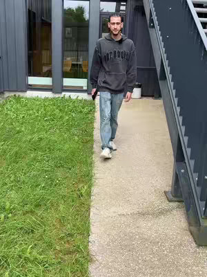 A man in a dark hoodie and jeans is walking towards the camera. The background includes a building with a glass window and a stairway. A patch of green grass is on the left and a walkway on the right.