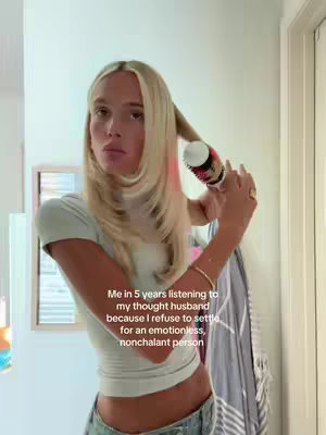 A woman with long blonde hair is styling her hair with a round brush. Her expression is neutral and she appears to be looking away. The scene is in a bathroom with a light color palette and a framed mirror in the background.