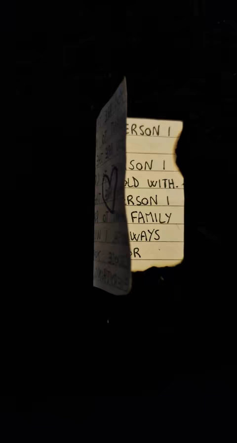 A crumpled piece of paper with handwritten text on it is centered in a dark composition, suggesting a note or message. The paper has a burnt edge on the right side, indicating it may have been exposed to fire. The left side is slightly obscured, suggesting a partial view of the full text. A heart appears in the middle. The background is completely black.