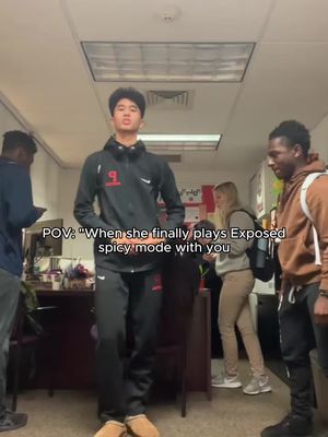 A full shot shows a young man in a black track suit standing in a hallway, with two other people in the background. The text overlay reads 'POV: 'When she finally plays Exposed spicy mode with you.'