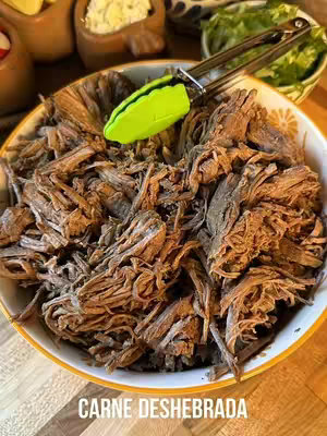 A close-up shot of a bowl filled with shredded meat, likely beef, with a pair of green tongs resting on top. The meat appears to be well-cooked and seasoned. The background includes other bowls and possibly ingredients, all on a wooden surface.