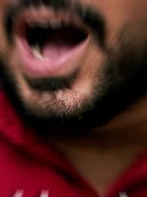 Close-up shot of a man's face, with a blurred, radial effect. His mouth is wide open, as if shouting or surprised, revealing his teeth and tongue. He has a beard, and is wearing a red top.