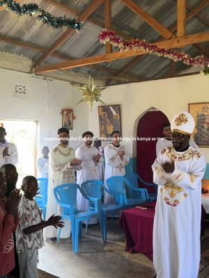 An indoor shot of a religious ceremony with multiple people in attendance. The room has a high ceiling with exposed wooden beams adorned with decorative garlands. A man in a white and gold ceremonial outfit stands at the right, facing the viewer. Several other men in white robes and some with white clerical outfits appear in the center of the image.