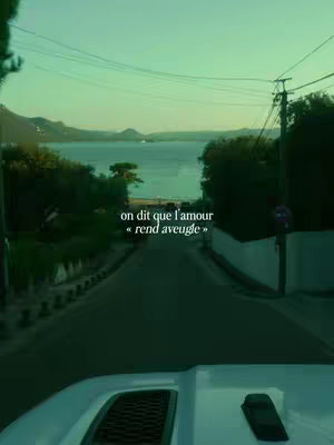 The image shows a view from a car, likely a white vehicle, on a road with sea in the background, with a text overlay. The text appears to be in French. The road is leading towards a body of water, surrounded by trees and greenery, with mountains visible in the distance. The sky has a light greenish tinge. The focus is on the road ahead.