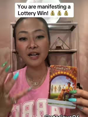 A selfie-style video cover with a woman holding a tarot card, overlayed by text. The woman is in the foreground, smiling and looking at the camera, with colorful nails. Behind her is shelving with a clear glass pyramid.