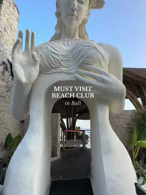 A white stone statue of a person with arms raised and hands open. The torso of the statue has an opening, through which a view of the beach and blue sky can be seen. The statue has a decorative headpiece, with a text overlay over the opening in the torso. 