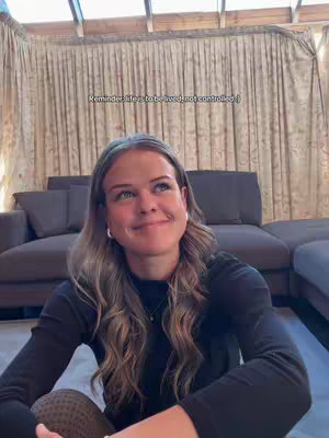 A woman with long, wavy hair sits in front of a large, neutral-toned couch, looking upward with a soft smile. The lighting is natural, and the background includes curtains and a glass ceiling, suggesting an interior space with ample natural light.