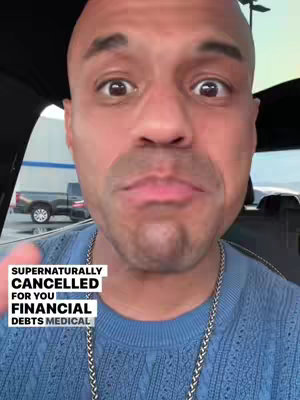 A man with a shaved head and a gold chain is looking directly at the camera with a surprised expression. He has his mouth slightly open. Overlay text in a box says 'Supernaturally Cancelled For You Financial Debts Medical'. The background is blurred, showing a car interior and part of a truck.