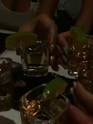 The image shows a close-up view of several hands holding shot glasses filled with a golden liquid, presumably alcohol. Each shot glass has a lime wedge resting on the rim. The lighting is dim, suggesting an indoor setting.