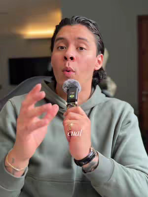 A man with slicked-back hair is holding a microphone and speaking, gesturing with his left hand. He wears a green hoodie and a watch with a black strap on his left wrist. A gold ring is visible on his ring finger. Text overlayed on image: 'cual'. The background is blurred but appears to be an interior setting.