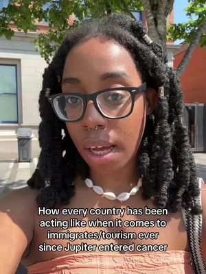 A selfie-style video shows a young woman with black locs, glasses and a seashell necklace, in front of a building. The text overlay adds context.