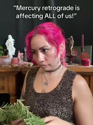 The image shows a person with vibrant pink hair and is positioned at a slight angle, looking down with a serious expression. The person wears a dark, patterned top and a choker necklace. They hold a bunch of green herbs in their hands, possibly rosemary. The background is a blurred mix of dark and wooden elements. Text reading "Mercury retrograde is affecting ALL of us!" is at the top.