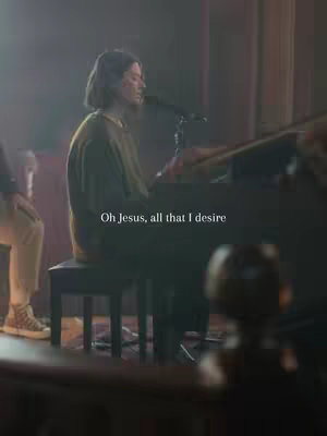 A woman plays a piano in a dimly lit setting, likely a church. The image is composed with a vertical layout, and the color palette is muted with natural light and shadows. Text overlay appears in white at the bottom.