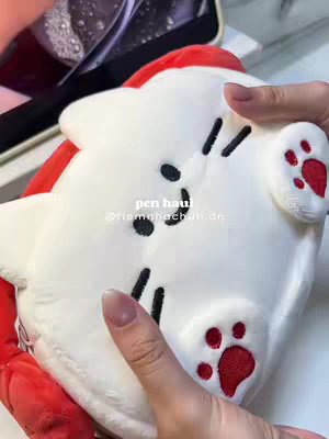 The cover image showcases a close-up view of a white plush toy shaped like a cat. The cat has black dots, lines, and a red paw print. A hand with manicured nails is seen holding the toy.