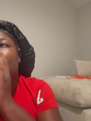 The image is a selfie featuring a person, likely a woman, in a close-up shot. She is seen from the side, with a black head covering and a red t-shirt. Her hand is raised to her face, partially obscuring her mouth. The background is a plain light gray wall and a portion of a beige couch, providing a simple setting. The overall composition is dark with some light.