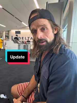 A man with long hair, a beard, and a backwards cap is seated in what appears to be an airport terminal, with a text overlay reading 'Update' superimposed on the image. He is wearing a dark blue button-down shirt and red and white striped shorts. The background shows the Delta check-in area.