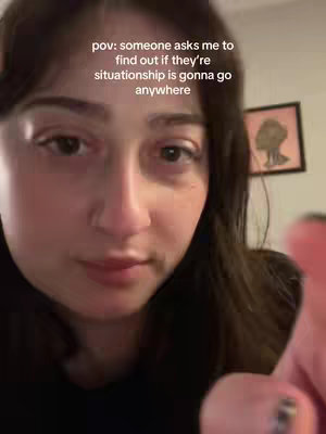 A selfie-style video cover image. The creator, a person with long, dark hair, is positioned in the center and slightly angled towards the lower-left of the frame. The face is the focal point. The creator is looking down, a hand partially visible in the bottom right corner. The background is softly blurred, revealing what appears to be a framed picture. A text overlay, written in white, is superimposed over the image, positioned in the upper-left corner.