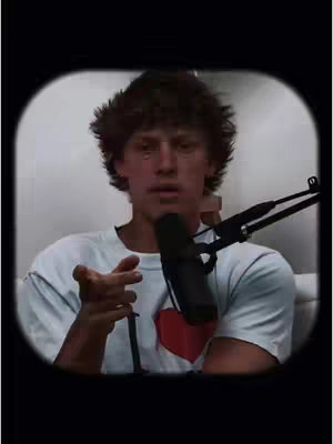 A young man with brown hair is centered in a circular frame. He is wearing a white t-shirt with a red heart graphic. He is holding a microphone and gesturing with his right hand facing the viewer. The background is a blurry light color.