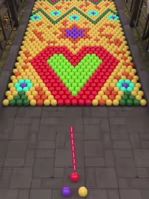 The image shows a top-down view of a game-like setup made of colorful balls, with a red line indicating the path of a ball towards a target. The setup features a large design of a heart, made of green and red balls situated amidst a pattern of yellow, orange, and turquoise balls with a dark grey surface.