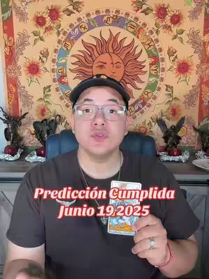 A medium shot shows a man holding a tarot card, text overlays appear and the background includes astrological decorations. Central to the image is the man, positioned in the foreground and gazing towards the viewer. He wears a black cap, glasses, and a dark-colored shirt. In his hands, he holds a tarot card. Overlayed on the image is bold red text. The background features a complex tapestry with a sun and radiating lines design. Behind the man are decorative elements like statues and a plate. The color palette uses warm tones like red, orange, and gold, giving the image vibrancy and depth.
