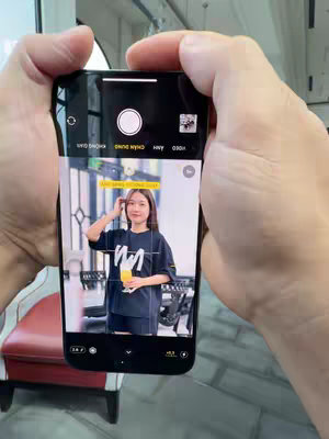 A person holds an iPhone displaying a camera interface focused on a woman. The woman is standing inside a room, wearing dark clothing and holding what appears to be a drink. The iPhone screen dominates the image, with elements of the camera UI visible, and two hands are seen gripping the phone.
