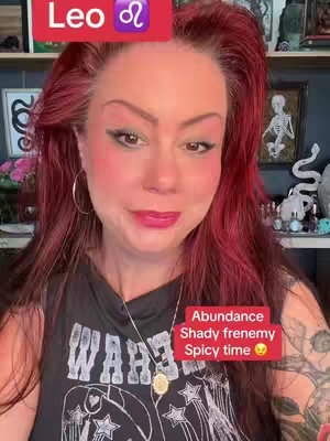 A woman with red hair and colorful makeup is centered in the frame, smiling and looking directly at the viewer. A red banner with text and a zodiac symbol is displayed on the top left. A text bubble containing information is in the lower right of the frame.