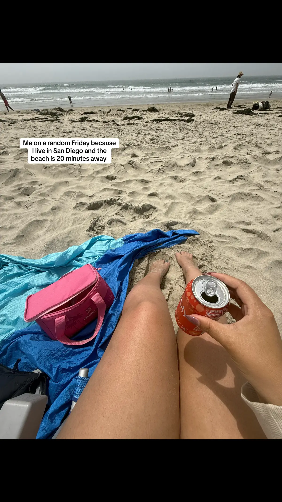 A person at the beach on a sunny day. The person's legs are in the foreground with a Coca-Cola can in their hand. There is a pink bag, a blue towel, and sand in the foreground as well. In the distant background, there is water and people. The image conveys relaxation and happiness.
