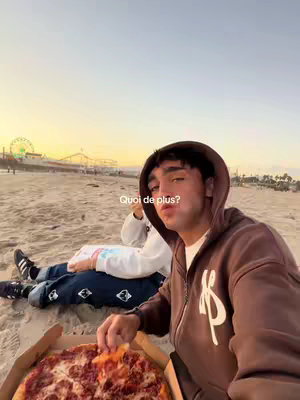 A young man wearing a brown hoodie is seated on a sandy beach at sunset, holding a slice of pizza. Behind him, another person is lying down. In the background, there's a ferris wheel and other amusement park structures visible, silhouetted against the sky.
