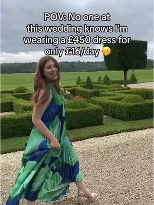 A woman in a colorful dress is walking towards the camera on a paved path. The background features a meticulously manicured garden with neatly trimmed hedges and trees under a cloudy sky.