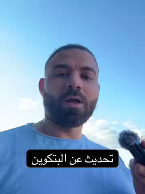 A man with a beard and short hair stands outdoors against a blue sky, holding a microphone. A black rectangle at the bottom of the image contains Arabic text.