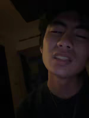 A close-up shot of a young man's face, taken in a dimly lit environment. His eyes are closed, and his face shows a slightly strained expression, as if he is speaking or reacting to something. The lighting is moody with shadows, highlighting his facial features.