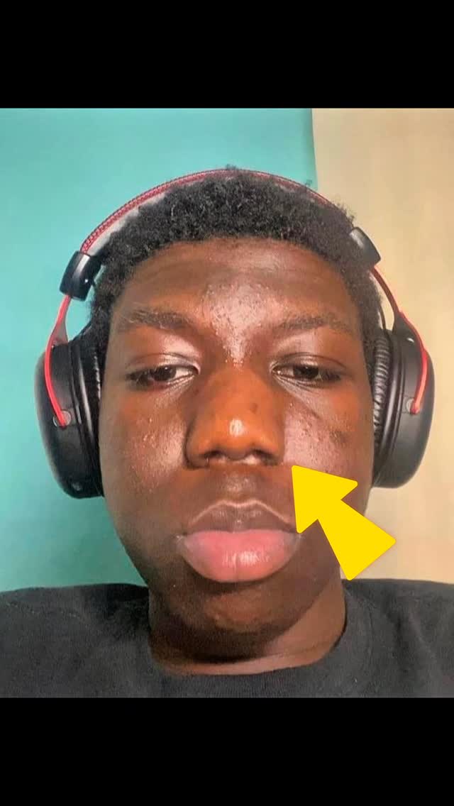 A close-up selfie of a young Black man with headphones. He has a focused expression with a slight pout. A yellow arrow points to his face.