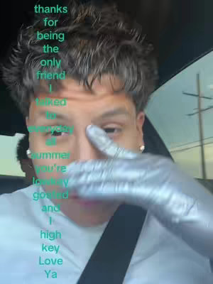 A selfie-style shot taken from inside a vehicle. The main focus is a young man, his face partially obscured by a gloved hand. Text overlays in a teal color are vertically stacked on the left, expressing a message of gratitude to a friend. The background subtly reveals the car’s interior and a glimpse outside.