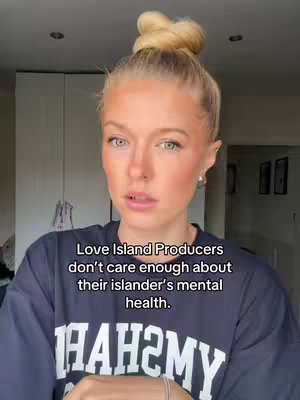 A young woman with blonde hair styled in a bun is looking at the camera with a concerned expression. She is wearing a dark blue shirt with a white graphic. The background is out of focus but appears to be a room with white cabinets and some framed pictures.